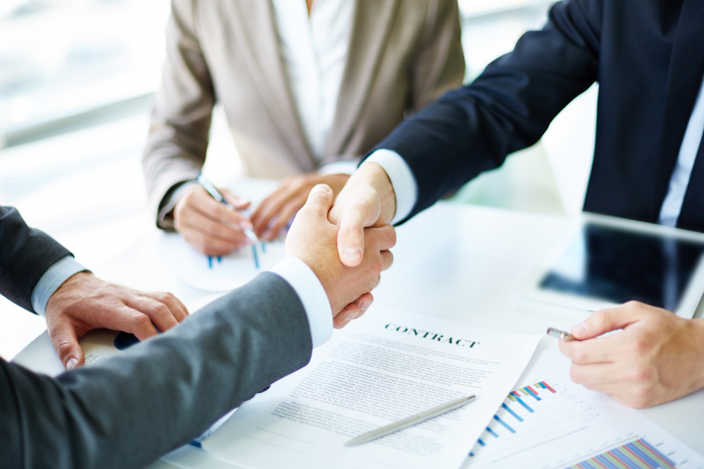 Revisão de contratos. Imagem: Dois homens de terno apertando as mãos com um contrato em cima da mesa.