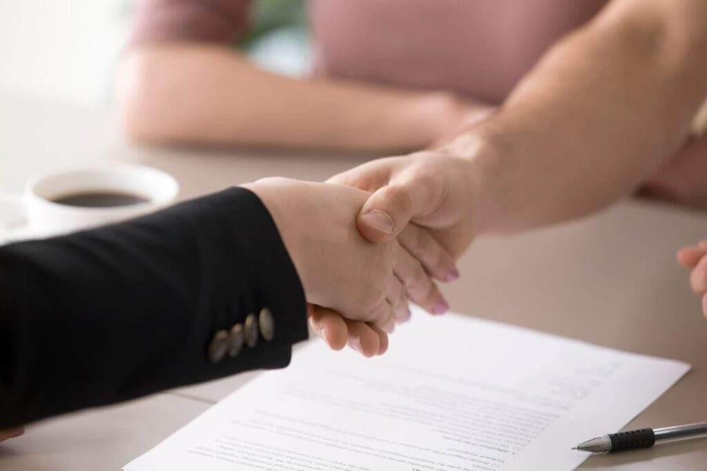 Imagem de duas pessoas apertando as mãos e, entre elas, está uma mesa com café e um dos tipos de contratos de trabalho.