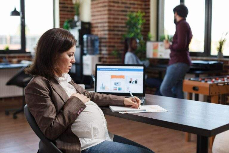 Imagem de mulher na mesa de escritório com um computador e fazendo anotações, com a mão na barriga, grávida, simbolizando a ideia de auxílio de um advogado especialista em licença-maternidade.