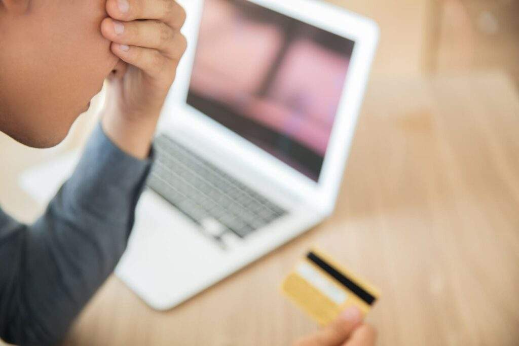 Homem com cara de preocupação, olhando para o cartão de crédito, com um notebook ao lado, simbolizando o arrependimento de compra.